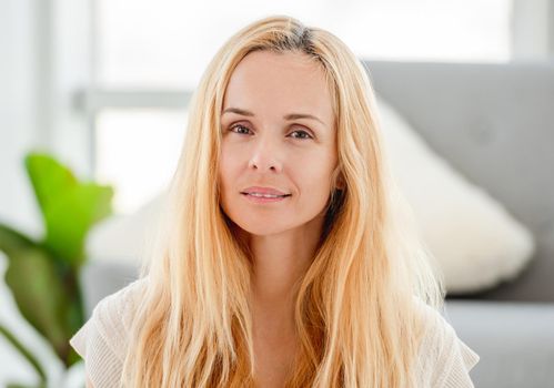 Beautiful young blond girl smiling and looking at the camera at home. Portrait of pretty woman with light blurred interior on background