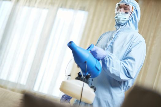 Man in protective blue suit holding microparticle gun for virus prevention in hotel room. Coronavirus and quarantine concept