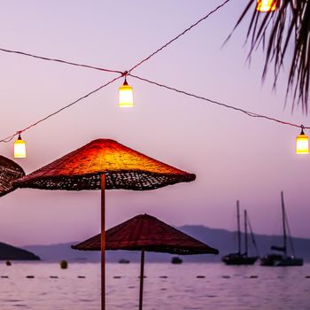 Beach umbrellas and lanterns against the backdrop of the sea, islands, mountains and yachts at sunset. Summer vacation concept