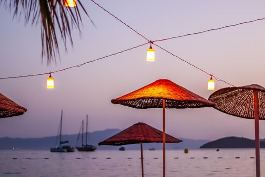 Beach umbrellas and lanterns against the backdrop of the sea, islands, mountains and yachts at sunset. Summer vacation concept