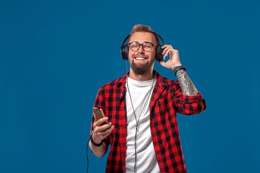 Happy young man listening to music with headphones. Handsome smiling guy in checkered shirt with closed eyes dancing with headphones. Isolated on blue background. Concept of people emotions
