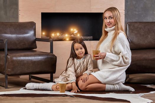 Young beautiful blonde woman hugs her little daughter on the couch with mugs of warm drink. Mother care about the child at home. The tenderness of my mother's embraces. Christmas winter comfort