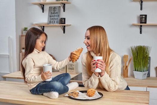 Shot of cheerful mother and daughter sit together at kitchen table, drink hot tea in morning, have pleasant friendly talk between each other. Curious girl asks something in mum during coffee break.