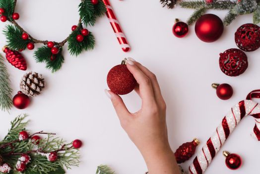 A female hand with a white manicure holds a red ball on a white background with Christmas branches and red decorations.