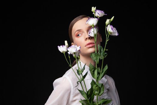 Pretty young woman in elegant white blouse touching face with natural flowers and looking away against black background