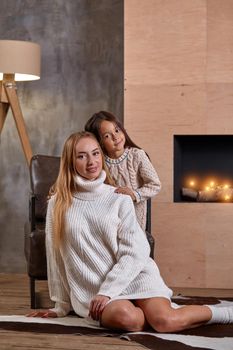 Mom with little girl in white sweaters relaxing on floor near a cozy sofa, Christmas lights in the fireplace