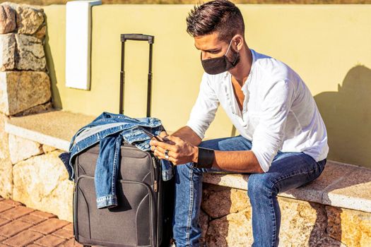 Solo traveller waiting for public transport sitting on bench wearing safe protective black face mask using smartphone to pass tedious waiting. Stylish guy with travel trolley bag in social distancing