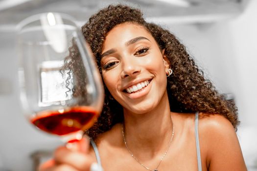 Close-up of young beautiful Afro-American woman toasting looking at camera in video conference POV simulation. New normal celebration online due to social distancing. Black Hispanic girl with red wine
