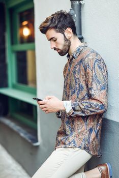 Young man wearing casual clothes looking at his smartphone in the street. Guy with beard and modern hairstyle in urban background