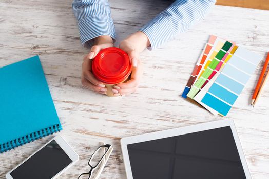 Office manager holding cardboard cup of coffee at wooden desk. Office workspace with tablet computer and accessories. Coffee break at office workplace. Young woman drinking coffee from disposable cup