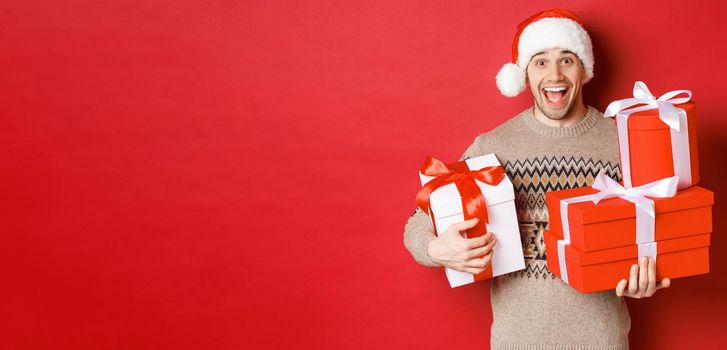 Concept of winter holidays, new year and celebration. Image of happy guy enjoying christmas, holding pile of presents and smiling amused, standing over red background.