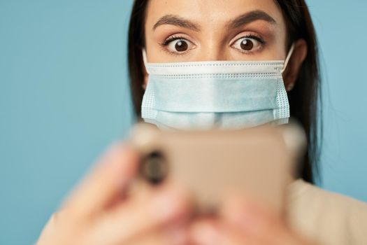 Surprised lady wearing protective mask and typing message on smartphone, isolated on blue background. Copy space. Quarantine, coronavirus concept