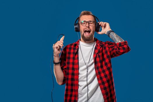 Dance Handsome young stylish man in headphones holding MP3 Player and dancing while standing against blue background. Emotional and people concept: young bearded man in checkered shirt. Hipster style. Copy space