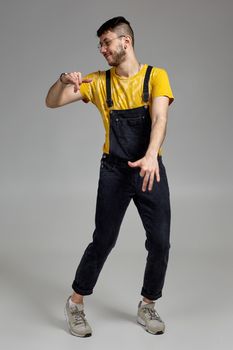 Full-length portrait of a smiling man in glasses, black jumpsuit, yellow t-shirt and gray sneakers fooling around in studio. Indoor photo of a man dancing on a gray background and looking away. Music and imagination.