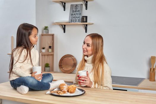 Shot of cheerful mother and daughter sit together at kitchen table, drink hot tea in morning, have pleasant friendly talk between each other. Curious girl asks something in mum during coffee break.