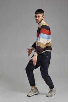 Full-length portrait of a handsome man guy in glasses, black jumpsuit, multi-colored sweater and gray sneakers fooling around in studio. Indoor photo of a man dancing on a gray background. Music and imagination concept.