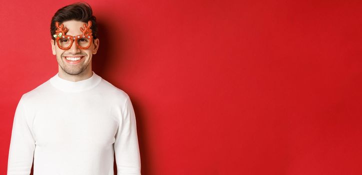 Close-up of attractive man in white sweater and party glasses, celebrating christmas, smiling amused, standing over red background.