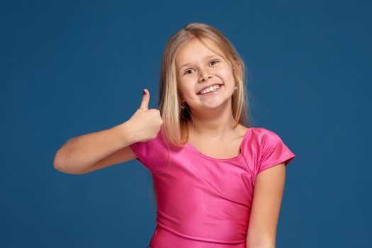 Close-up emotional portrait of caucasian little girl in pink shirt on blue background, making thumbs up gesture, looks happy