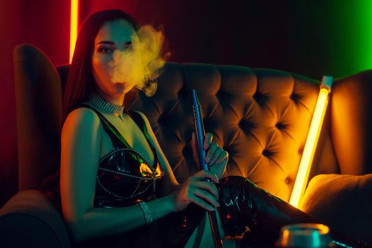 Lovely brunette girl with a perfect make-up, in a sparkling necklace, leather dress and patent boots is smoking a hookah exhaling a smoke sitting on an arm-chair at a luxury night club. Concept of pleasant spending time. Dark background with colorful neon backlights.