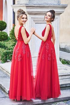 Two beautiful bridesmaids blonde and brunette ladies wearing elegant full length red chiffon bridesmaid dress with lace and holding flower bouquets. European old town location for wedding day.