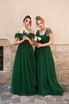 Two beautiful bridesmaids girls blonde and brunette ladies wearing elegant full length off-the-shoulder green chiffon bridesmaid dress and holding flower bouquets. European old town location for wedding day.