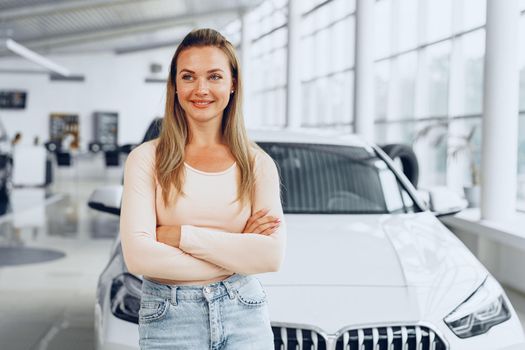Pretty happy young woman standing near new car in car showroom