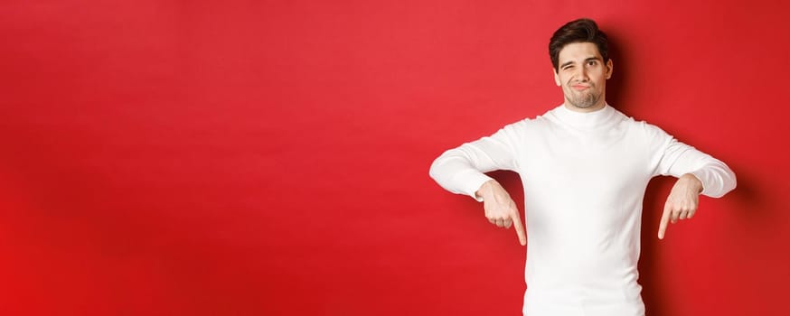 Portrait of attractive, skeptical young man in white sweater, grimacing and pointing fingers down, disagree and dislike something, standing over red background.