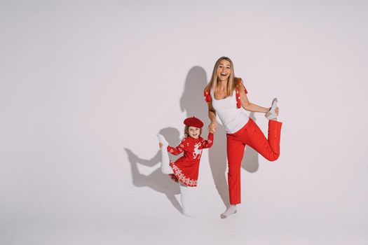 Studio portrait of cheerful mom and daughter in winter red dress with reindeer and mom in red trousers posing with leg bent behind.