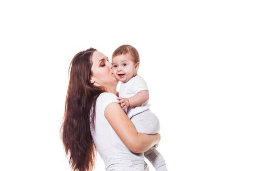Beautiful young mother holds her child in her arms and kisses her baby, wearing white cloths, on a white background