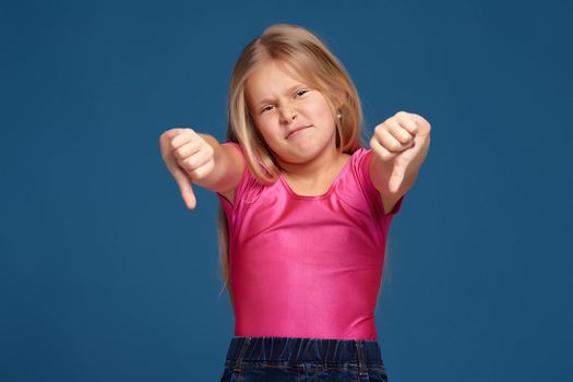 Close-up emotional portrait of caucasian little girl in pink shirt on blue background, making dislike, thumbs down gesture, looks unhappy
