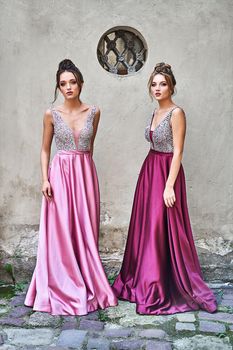 Two beautiful bridesmaids girls blonde and brunette ladies wearing elegant full length purple violet lilac lavender satin bridesmaid dress with silver sequined camisole top. European old town location for wedding day.