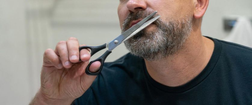 Portrait of Middle-aged handsome man cutting his beard with scissors. Selfcare during quarantine