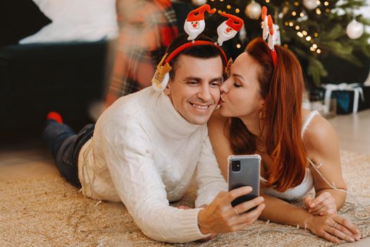 a happy married couple is lying on the floor at home near the Christmas tree and taking pictures of themselves.