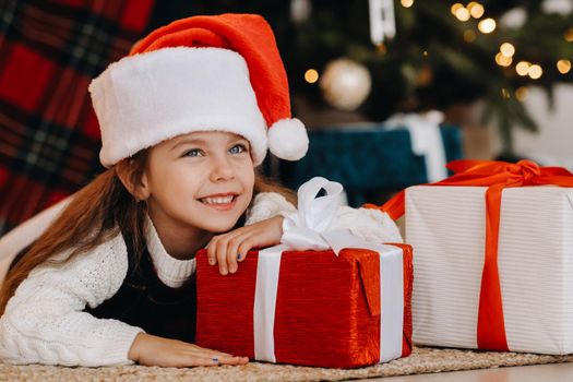 A happy little girl in a Santa Claus hat smiles with gifts in her hands.