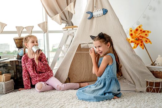 Little sisters playing with homemade telephone made of can and thread in room with wigwam