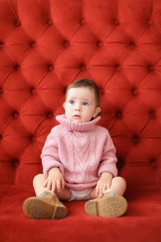 Little beautiful girl wearing pink sweater sitting on sofa with red background. Concept of baby photo session.