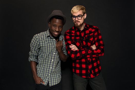 Studio shot of two stylish young men having fun. Handsome bearded hipster in a shirt in a cage standing next to his African-American friend in hat against a dark background. International friendship concept.