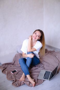 Young woman sitting near laptop and smartphone on bed, wearing white t shirt and jeans. Concept of modern technology and youth.