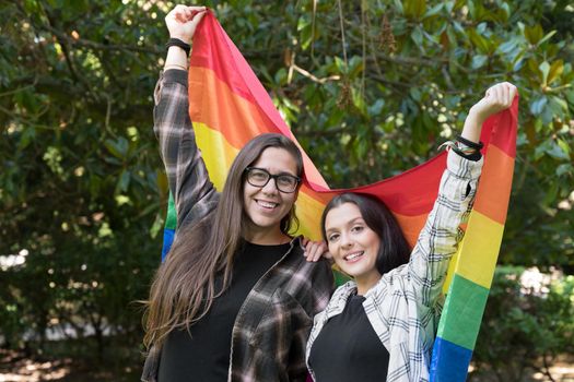 Lesbian couple from the lgtb community walking in the park with the rainbow flag while hugging, enjoying a sunny day. High quality photo.