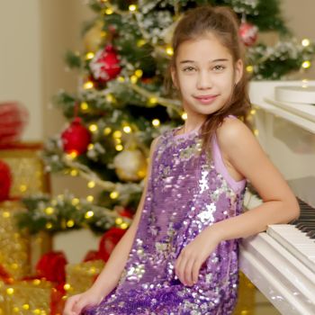 A cute little girl sitting near a white piano and a Christmas tree. The concept of the New Year, family holidays.