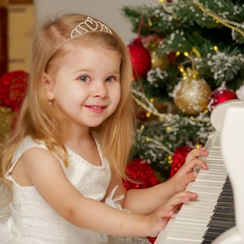 A cute little girl sitting near a white piano and a Christmas tree. The concept of the New Year, family holidays.