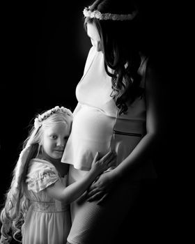 A little girl in the image of an angel hugs the belly of her eldest pregnant sister. The concept of family happiness, the birth of a child.