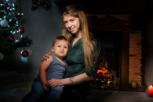 A young mother with a small son near the fireplace and the Christmas tree on the eve of the holidays. The concept of a happy family.