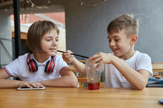 boy and girl sit outdoors at veranda of cafe. little caucasian schoolchildren sitting at table, playing with berries in cup of lemonade. childhood concept, fun, joy