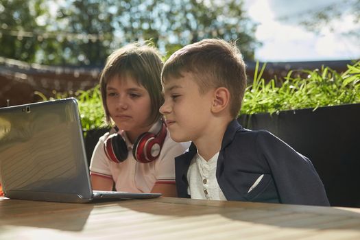 boy and girl sit outdoors, watch at screen of laptop on sunny day. little caucasian schoolchildren sitting at table at veranda of cafe, studying with computer, doing homework. online distant learning
