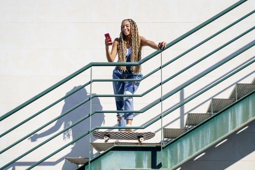 Young black female with coloured braids, looking at her smartphone with her feet resting on a skateboard.