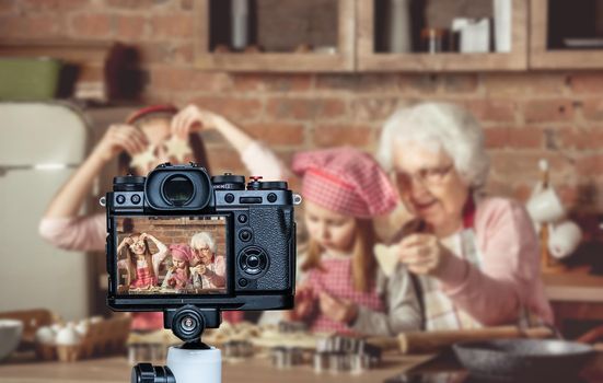 Grandma teaching granddaughters to bake homemade cookies. Little girl holding two star-shaped cookies in front of her eyes and smiling. Blogger profession. Shooting with a camera for a blog