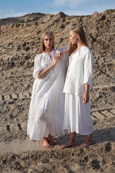 two young pretty twins with long blond hair posing at sand quarry in elegant white dress, skirt, jacket. stylish fashion photoshoot, summer photosession. identical sisters spend time together outdoors