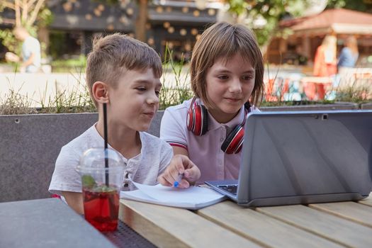 boy and girl sit outdoors, watch at screen of laptop. little caucasian schoolchildren sitting at table at veranda of cafe, studying with computer, watching movie. distant learning, online education