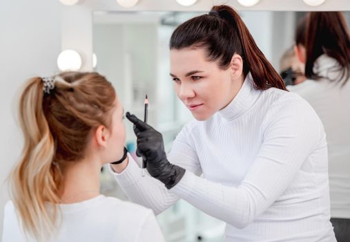 Permanent tattoo makeup master girl defines eyebrow shape with professional tools during microblading work process in beauty salon